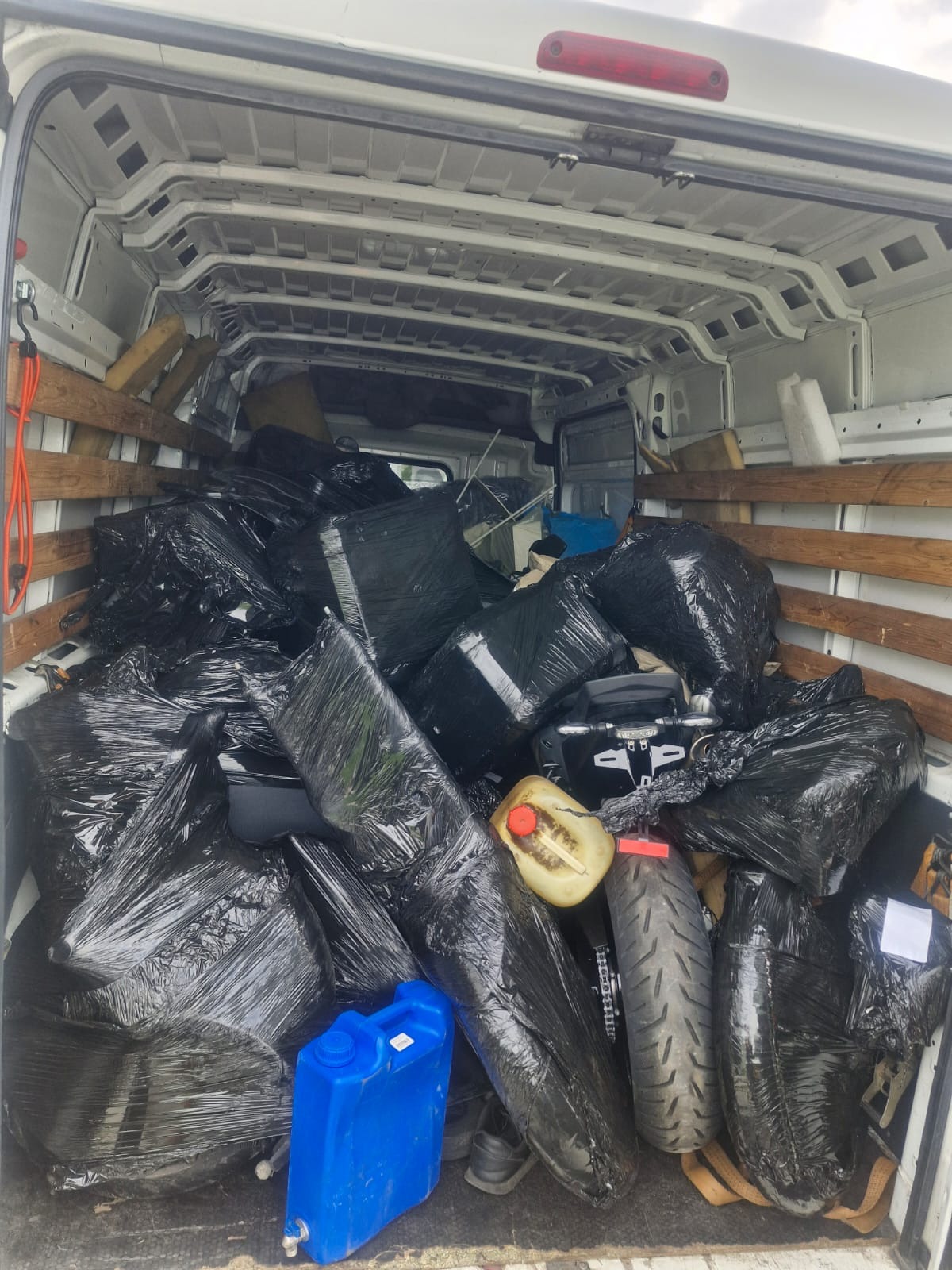 Wnętrze białego busa wypełnione po brzegi czarnymi workami, zabezpieczonymi folią, z widocznym niebieskim kanistrem i fragmentami motocykla.