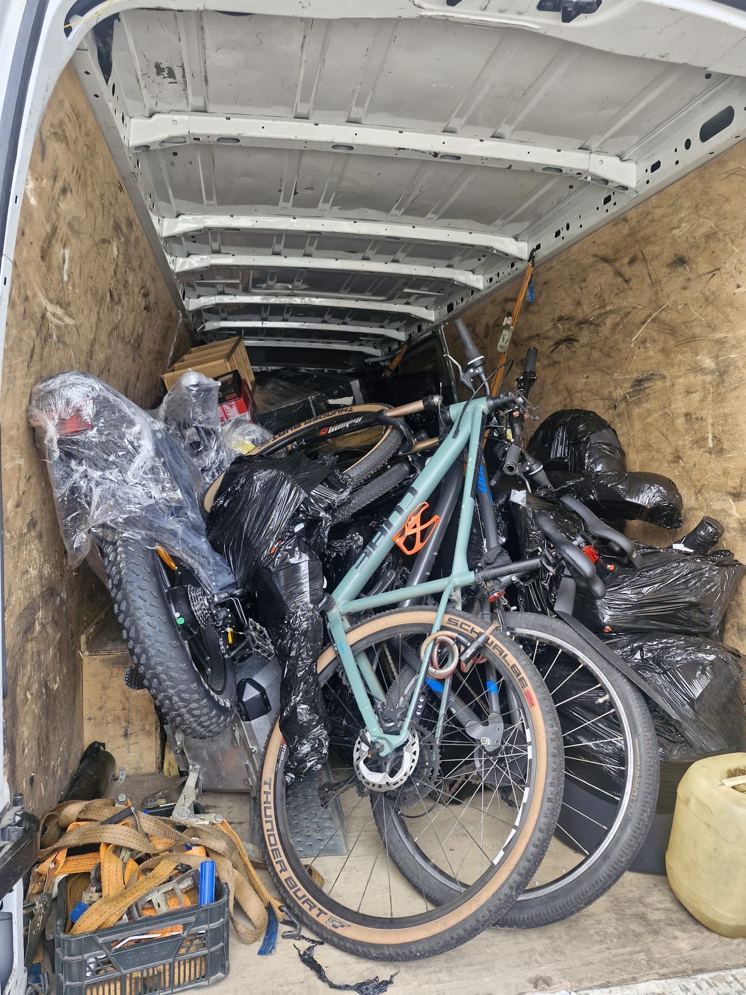 Wnętrze furgonetki wypełnione rowerami, niektóre owinięte folią i czarnymi workami, zabezpieczone pasami transportowymi. Widoczne drewniane ściany i biały sufit pojazdu.