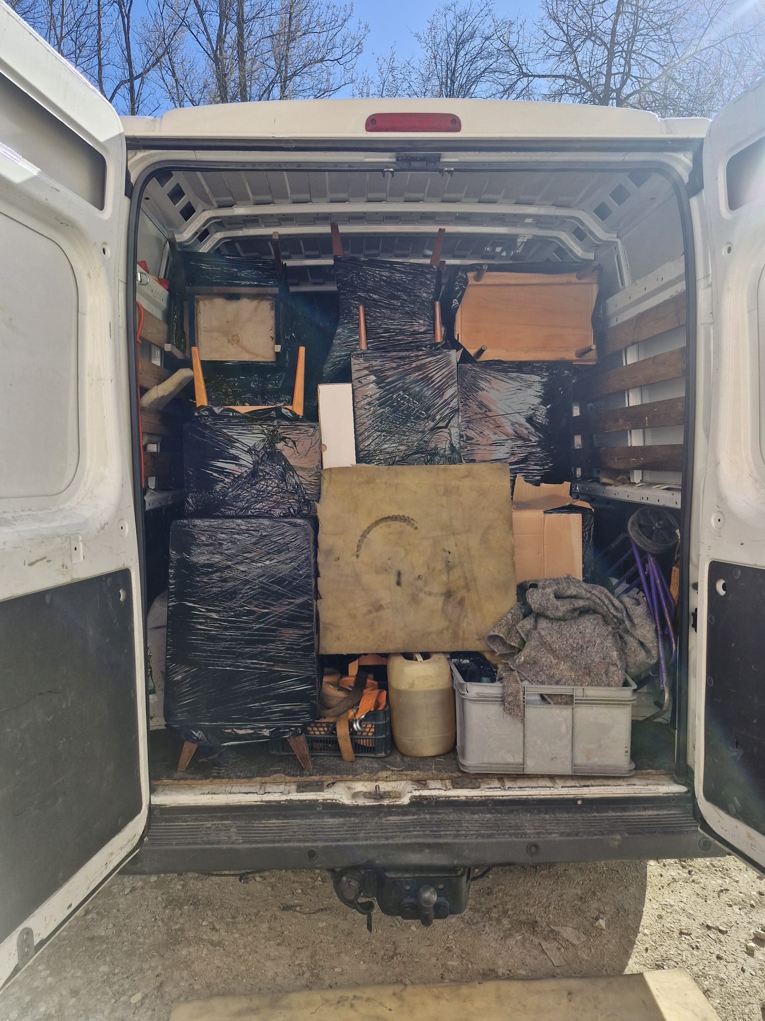 Wnętrze białego busa z otwartymi drzwiami, wypełnione meblami zabezpieczonymi folią i kocami, gotowymi do transportu. Widoczne krzesła, stolik i kartony.