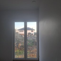 Ihor Serhiiev - Wnętrze w trakcie wykańczania: widok przez białe, dwuskrzydłowe okno na dom na wzgórzu. Jasne ściany, drewnopodobna podłoga. Minimalistyczny design, surowy stan.