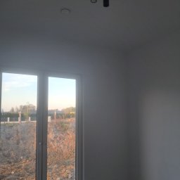 Ihor Serhiiev - Wnętrze pokoju z oknem i zamontowanym oświetleniem sufitowym. Widoczne przygotowania do prac wykończeniowych, z elementami materiałów budowlanych na podłodze.