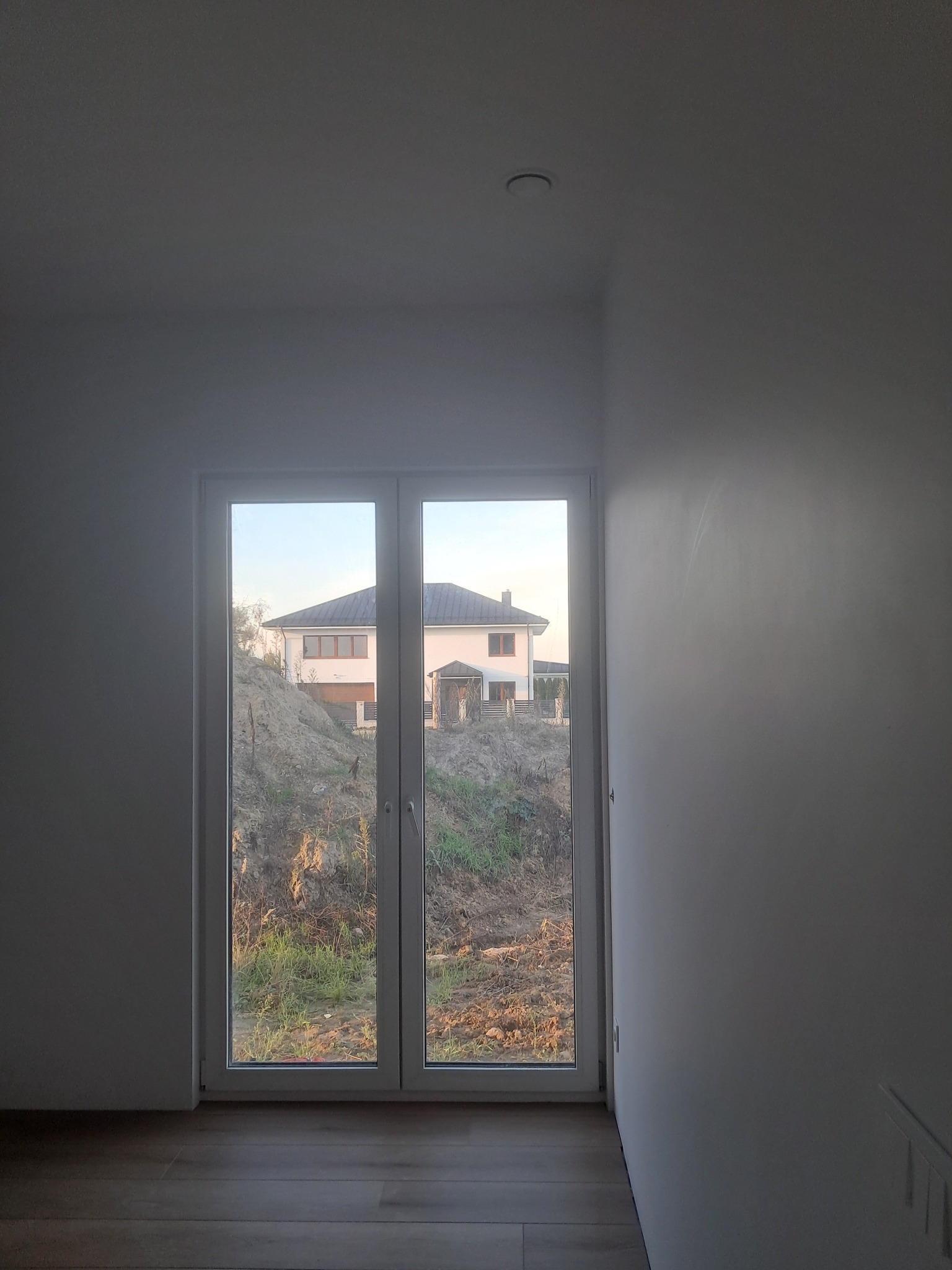 Wnętrze w trakcie wykańczania: widok przez białe, dwuskrzydłowe okno na dom na wzgórzu. Jasne ściany, drewnopodobna podłoga. Minimalistyczny design, surowy stan.