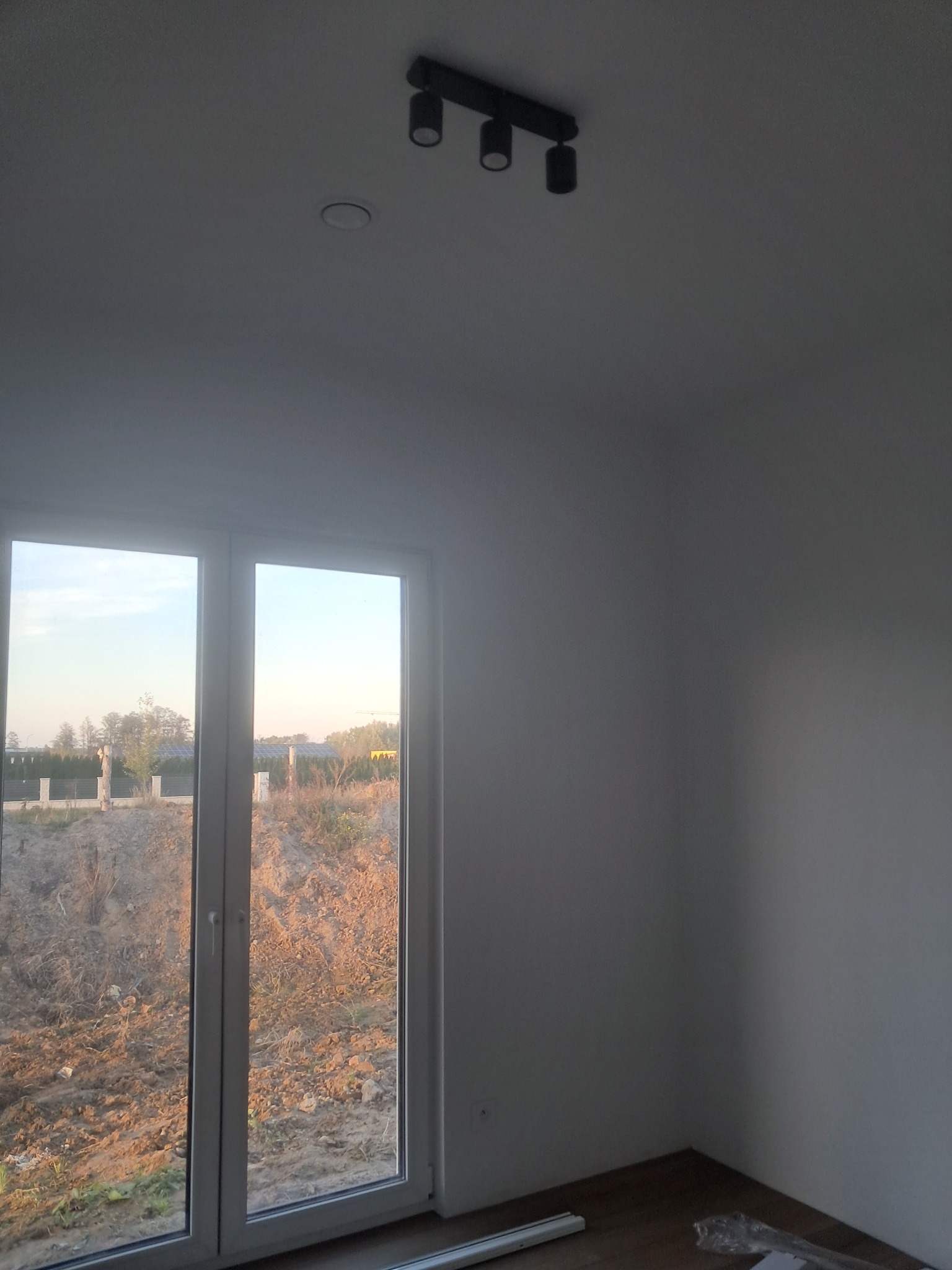 Wnętrze pokoju z oknem i zamontowanym oświetleniem sufitowym. Widoczne przygotowania do prac wykończeniowych, z elementami materiałów budowlanych na podłodze.