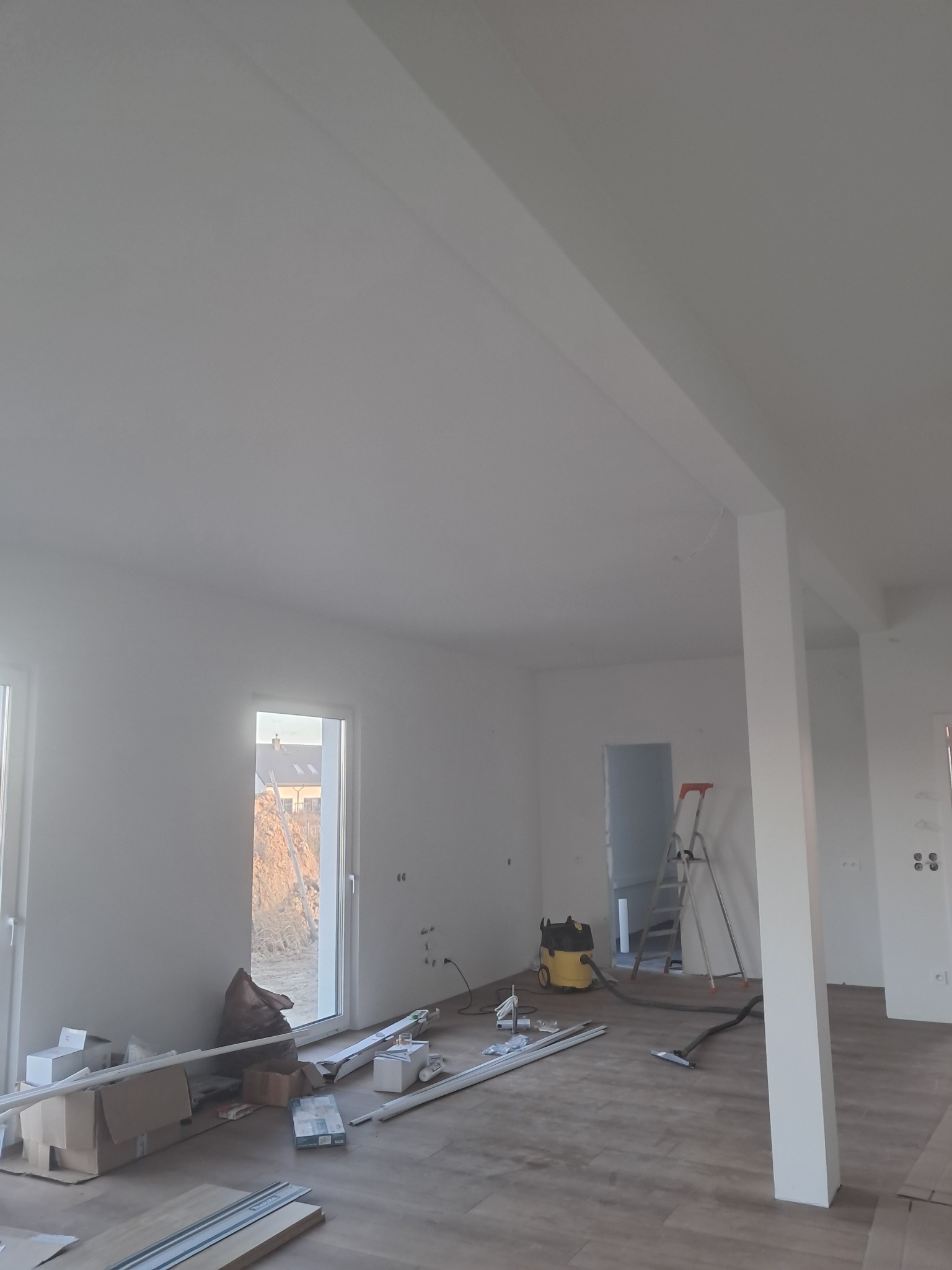 Jasne, przestronne wnętrze w trakcie prac wykończeniowych. Widoczne narzędzia, drabina, podłoga imitująca drewno i świeżo pomalowane ściany. Częściowo widoczne okno.