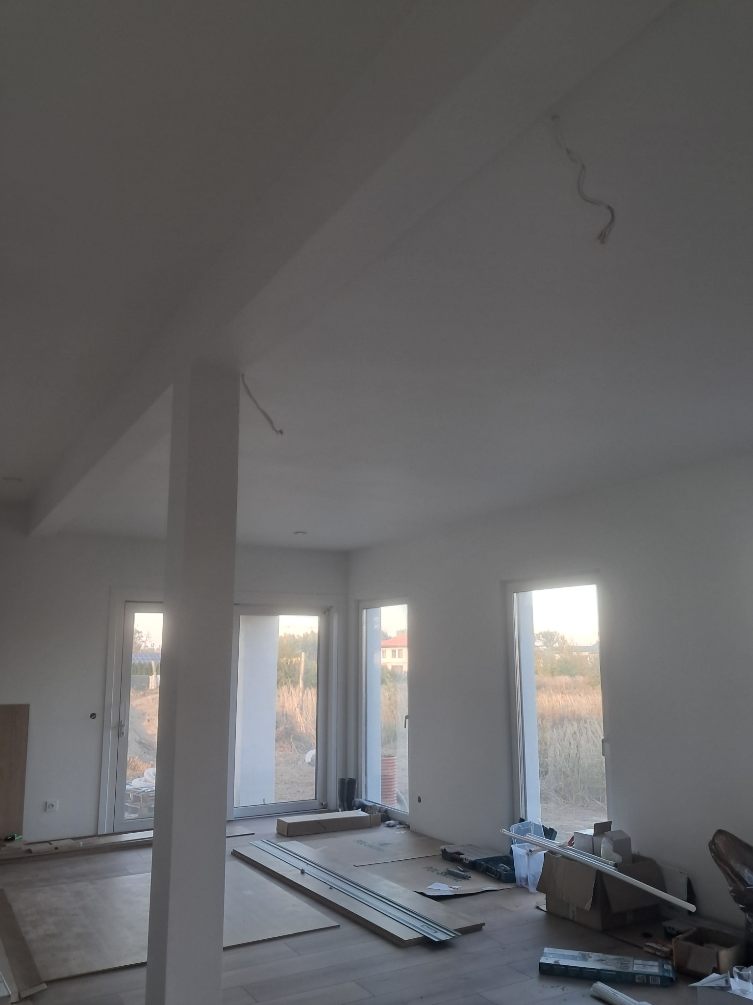 Jasne wnętrze w trakcie wykończenia: białe ściany, podłoga z paneli, okna z widokiem na zewnątrz. Widoczne materiały budowlane i narzędzia, sugerujące prace wykończeniowe.