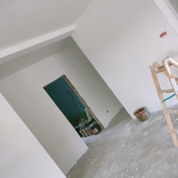 Lewy - Wnętrze po szpachlowaniu: gładkie, białe ściany, surowy beton na podłodze, drabina malarska i wiadro farby. Widoczny otwór drzwiowy z surową cegłą.