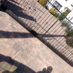 Lewy - Ukończony podjazd z kostki brukowej w odcieniach brązu, z dekoracyjną, kutą bramą w tle, częściowo zacieniony. Widoczny fragment domu i zieleni.