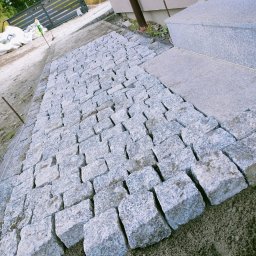 Lewy - Ścieżka z kostki granitowej w trakcie układania, widoczne surowe podłoże i fragment schodów z tego samego materiału. W tle ogrodzenie panelowe.