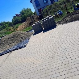 Lewy - Ułożona szara kostka brukowa na posesji, z przygotowanym podłożem i stosem kostki w tle, w słoneczny dzień. Widoczny fragment budynku i zieleń.