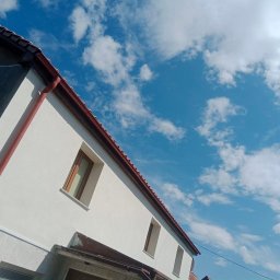 Lewy - Biała fasada domu z brązowymi oknami i rynnami na tle błękitnego nieba z chmurami. Część elewacji w trakcie prac wykończeniowych.