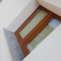 Lewy - Okno z drewnianą ramą i granitowym parapetem w białej, teksturowanej ścianie budynku, widziane pod kątem z dołu.