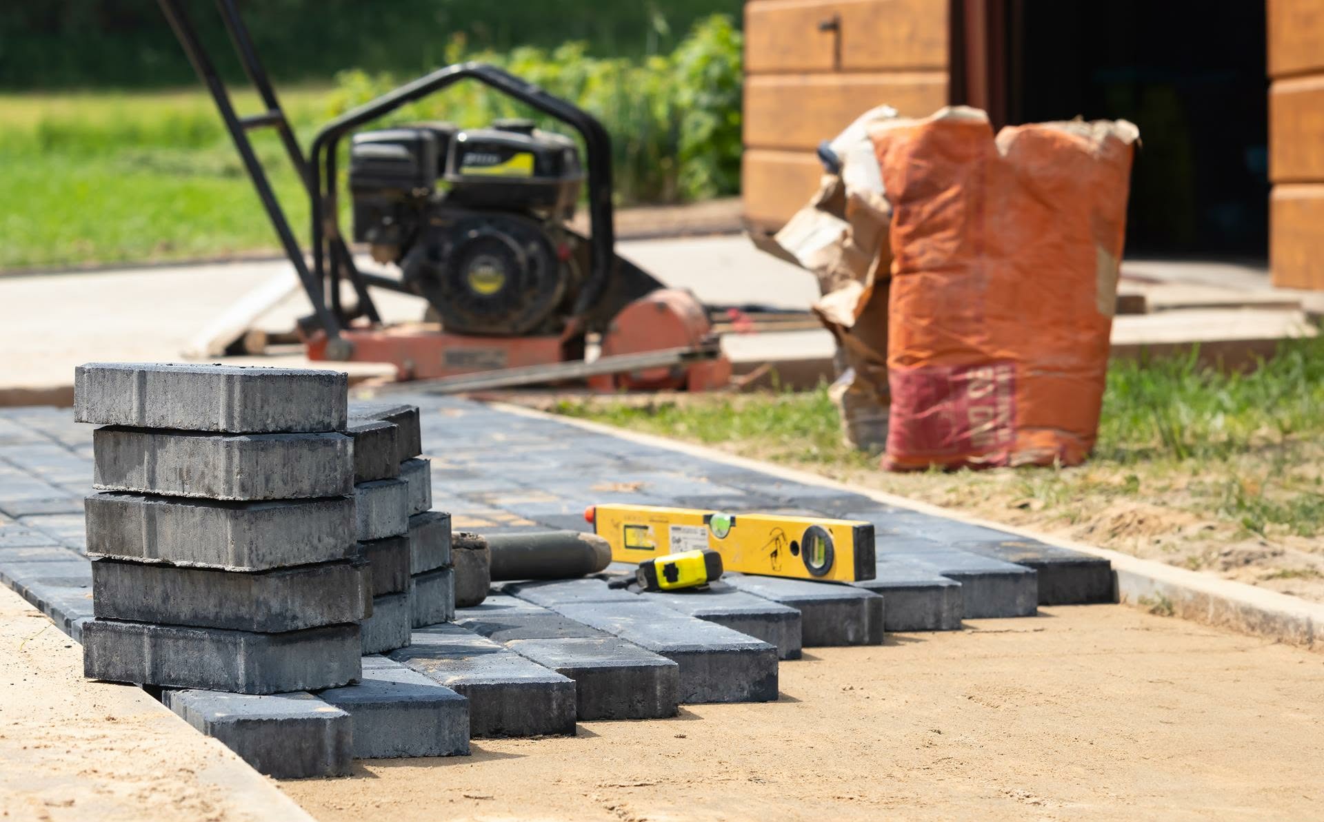 Częściowo ułożona, szara kostka brukowa z poziomnicą i zagęszczarką w tle, na przygotowanym podłożu z piasku. Widoczny fragment budynku i worki z materiałem.