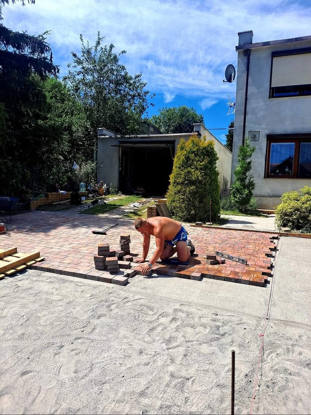Mężczyzna w trakcie układania czerwonej kostki brukowej na piaskowym podłożu. Częściowo ułożona nawierzchnia, widoczne narzędzia i budynek w tle.