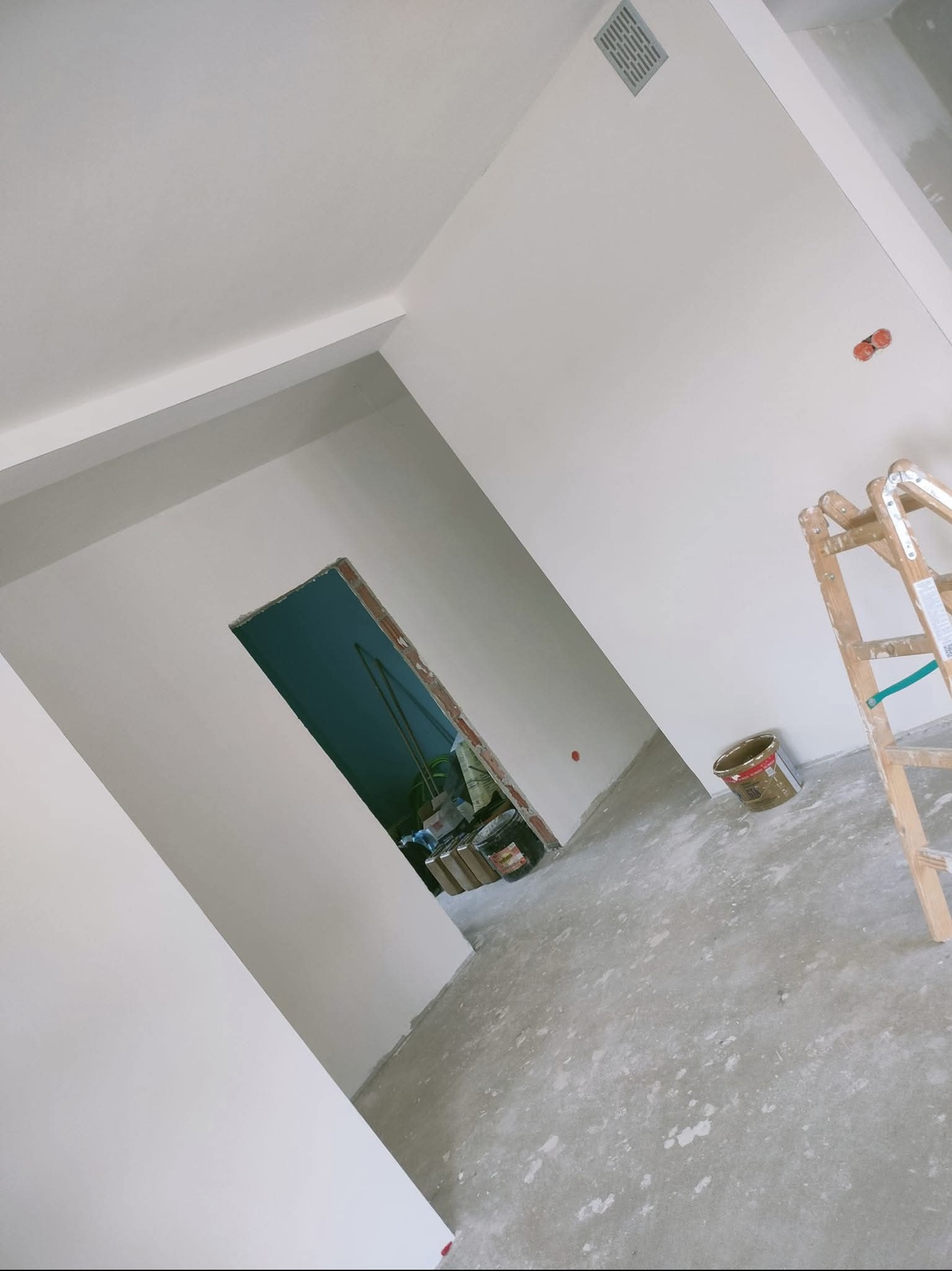 Wnętrze po szpachlowaniu: gładkie, białe ściany, surowy beton na podłodze, drabina malarska i wiadro farby. Widoczny otwór drzwiowy z surową cegłą.