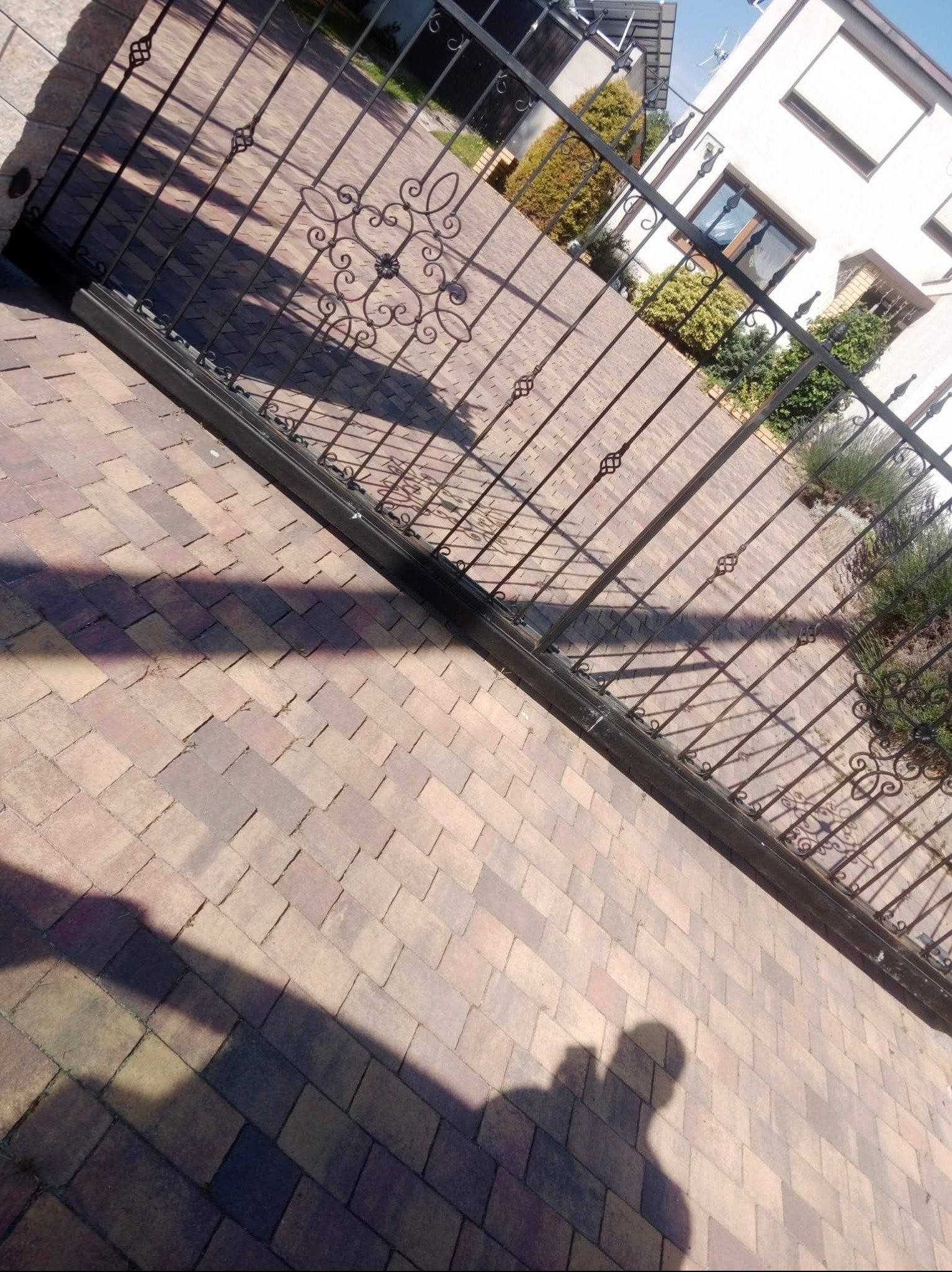 Ukończony podjazd z kostki brukowej w odcieniach brązu, z dekoracyjną, kutą bramą w tle, częściowo zacieniony. Widoczny fragment domu i zieleni.