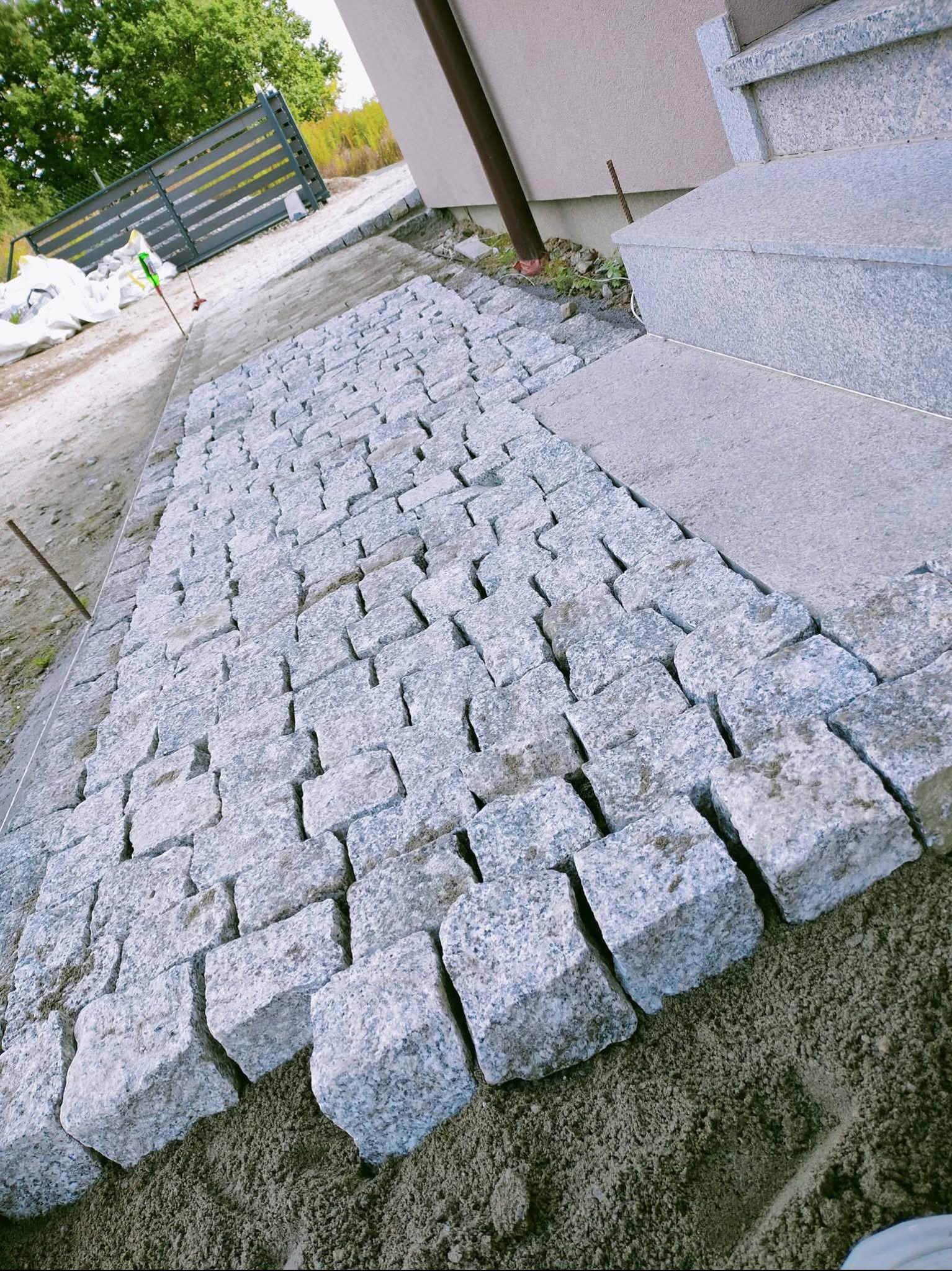 Ścieżka z kostki granitowej w trakcie układania, widoczne surowe podłoże i fragment schodów z tego samego materiału. W tle ogrodzenie panelowe.