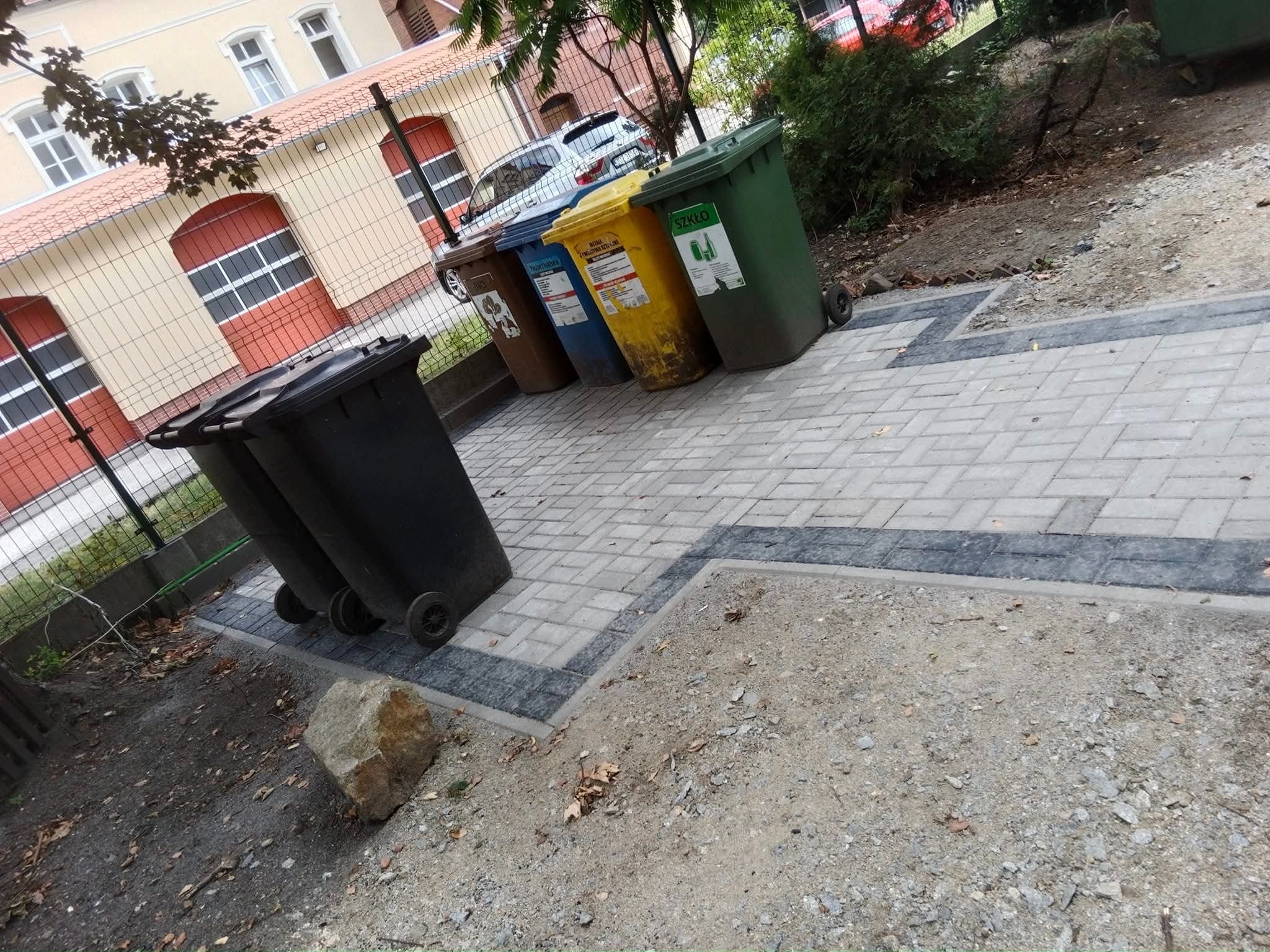 Ułożona kostka brukowa w szarych odcieniach, obramowana ciemniejszym pasem. W tle kolorowe pojemniki na śmieci i budynek z czerwonymi elementami.