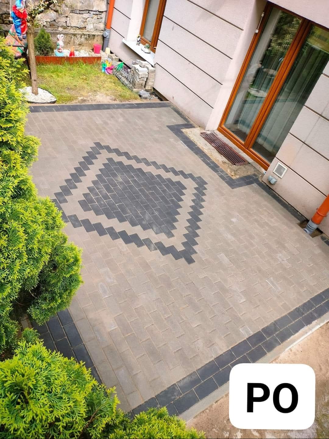 Ułożona kostka brukowa w geometryczny wzór na podwórku, obrzeża z ciemnej kostki, widok z góry, tuja po lewej stronie, budynek w tle.