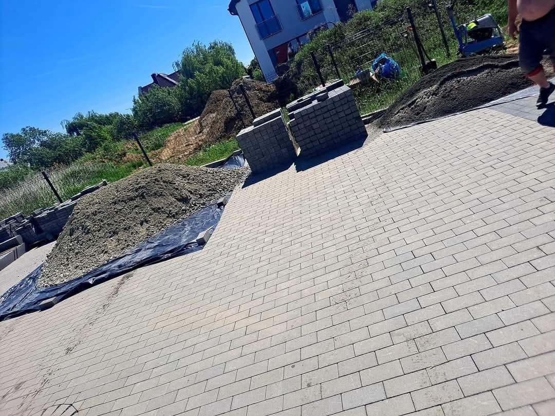 Ułożona szara kostka brukowa na posesji, z przygotowanym podłożem i stosem kostki w tle, w słoneczny dzień. Widoczny fragment budynku i zieleń.