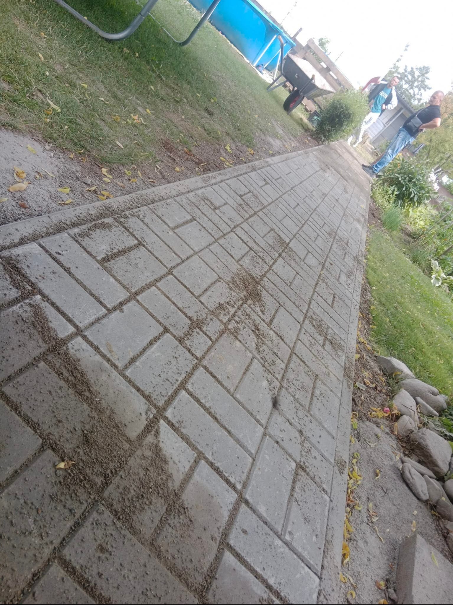 Ułożona ścieżka z kostki brukowej w ogrodzie, z widoczną trawą i elementami zieleni. W tle osoby i taczka. Perspektywa z niskiego kąta.