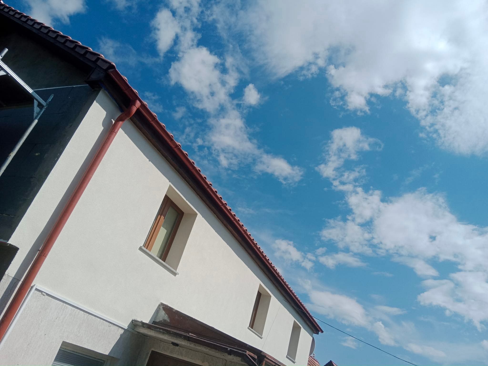 Biała fasada domu z brązowymi oknami i rynnami na tle błękitnego nieba z chmurami. Część elewacji w trakcie prac wykończeniowych.