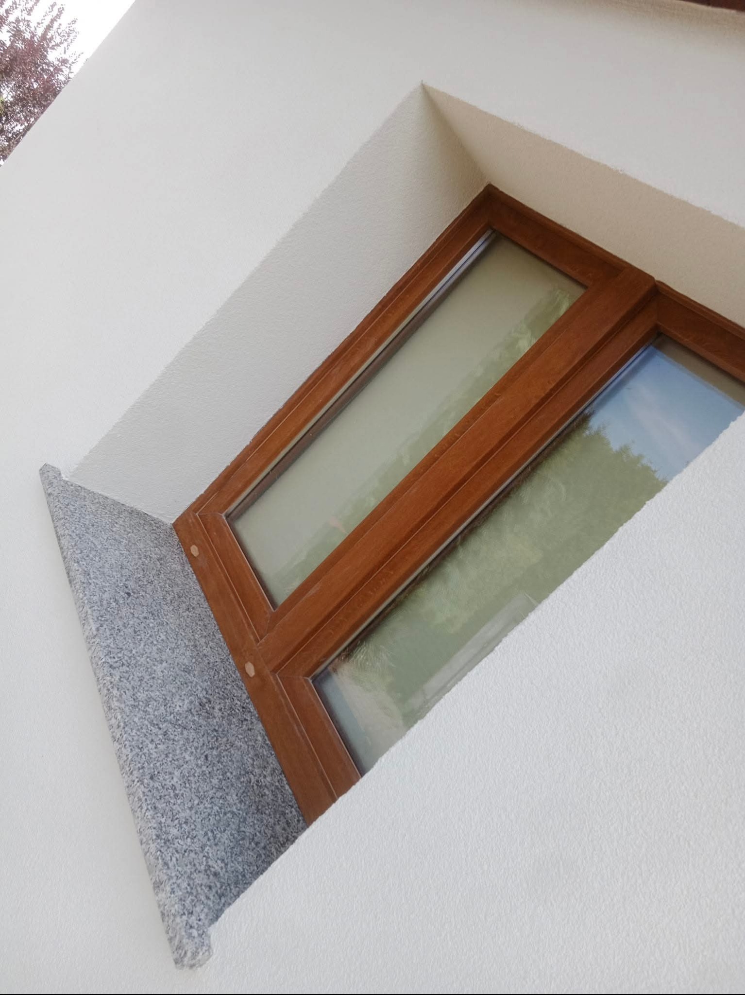 Okno z drewnianą ramą i granitowym parapetem w białej, teksturowanej ścianie budynku, widziane pod kątem z dołu.