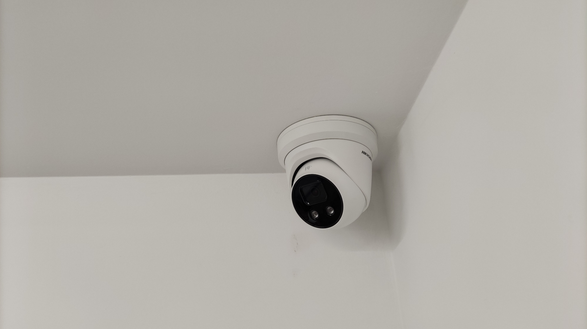 Kamera przemysłowa Hikvision, kopułkowa, zamontowana na suficie w rogu pomieszczenia. Instalacja monitoringu wizyjnego w Godowie. Białe ściany i sufit.