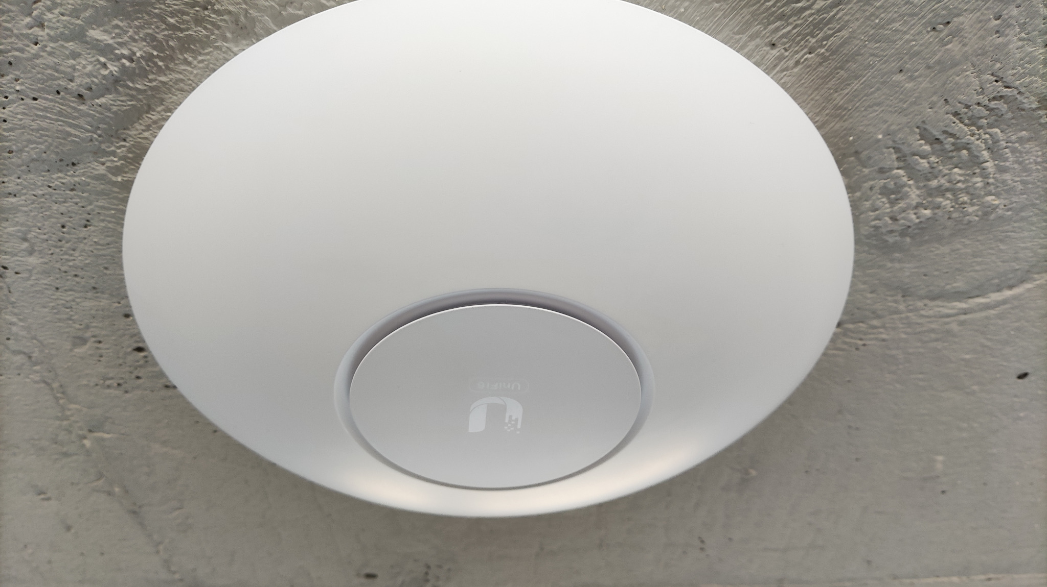 Zamontowany na szorstkim, betonowym suficie biały punkt dostępowy UniFi, widoczny z bliska, z subtelnym logo producenta na środku.