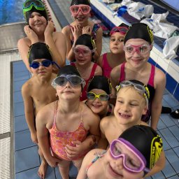 Uczniowski Klub Pływacki Wodnik Włocławek - Grupa uśmiechniętych dzieci w czepkach i okularach pływackich, stojących na kafelkach przy basenie, gotowych do zajęć. Widok z góry, jasne oświetlenie.