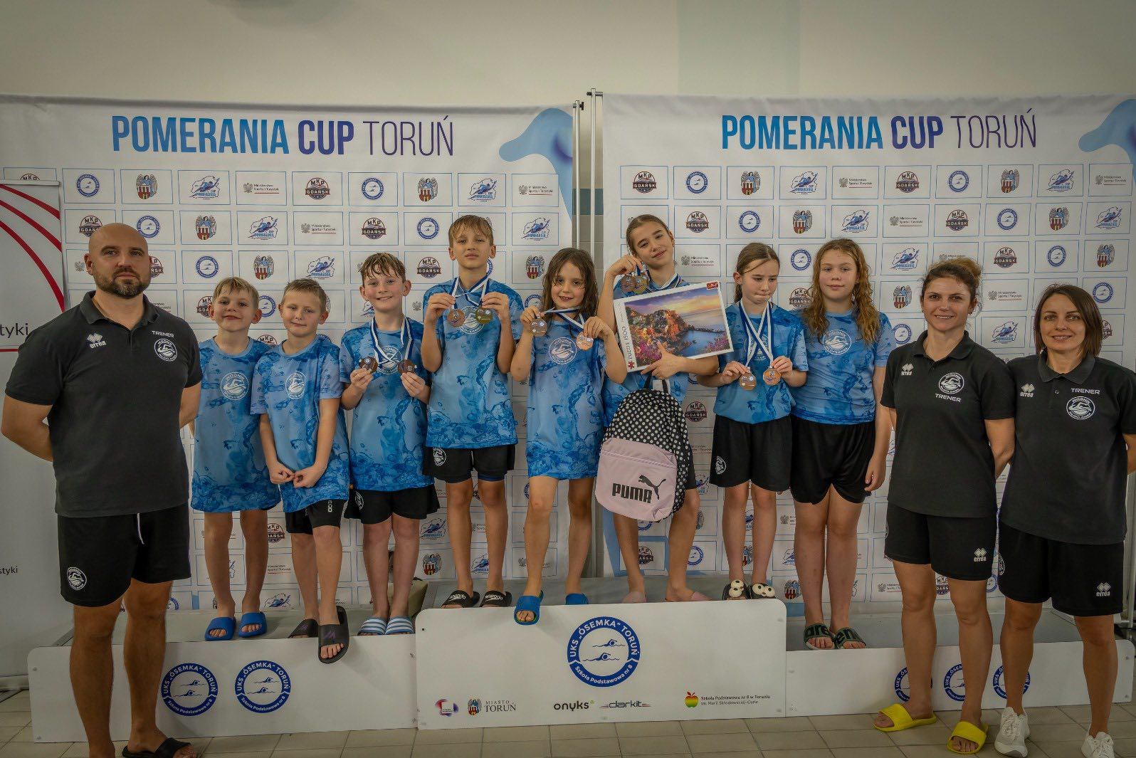 Grupa dzieci z medalami i trenerami na podium po zawodach pływackich Pomerania Cup Toruń. Uśmiechnięte twarze, sportowa atmosfera, widoczny baner z nazwą zawodów.