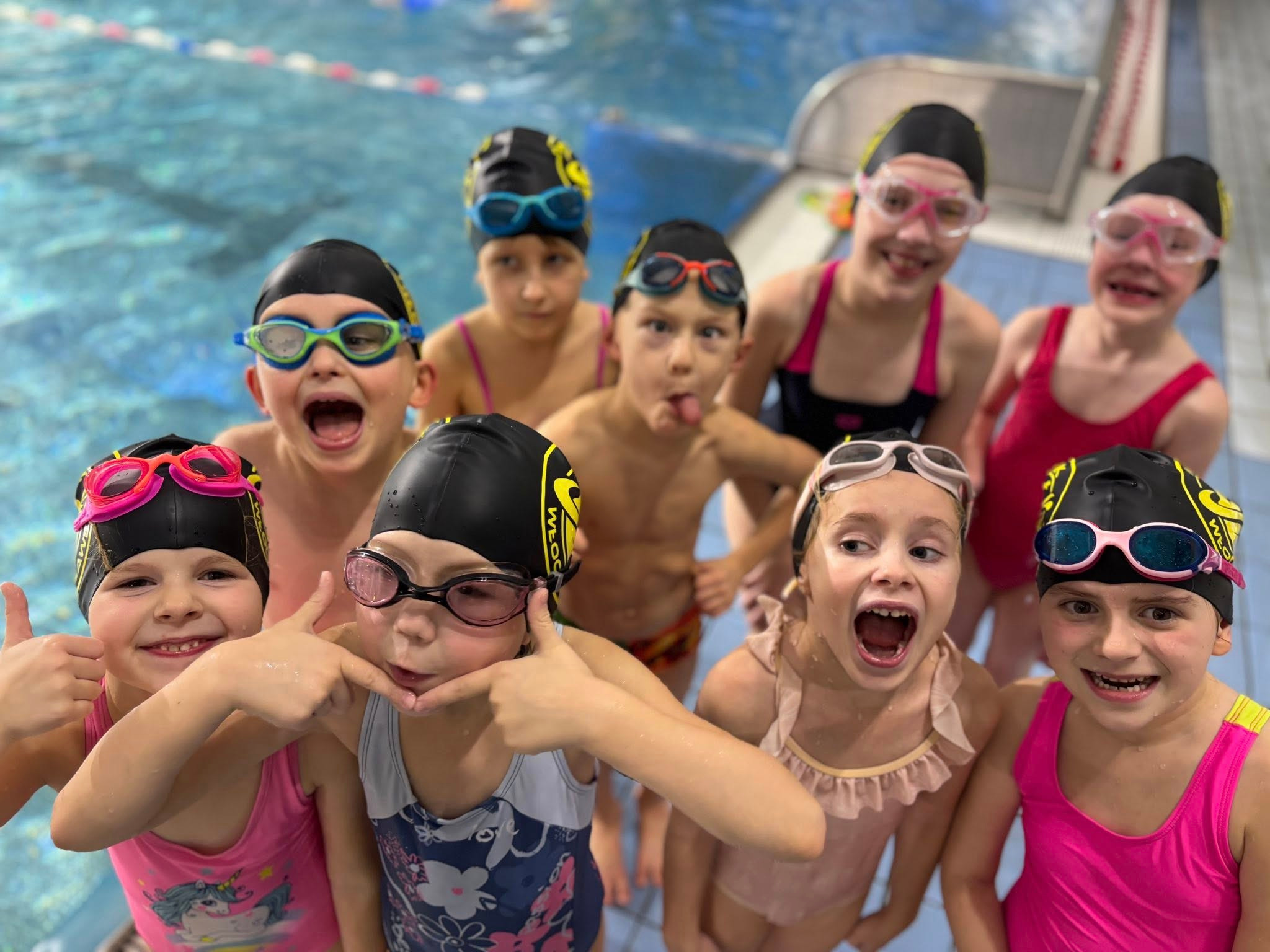 Grupa roześmianych dzieci w czepkach i okularach pływackich pozuje na tle basenu. Radosne miny i gesty, wesoła atmosfera zajęć pływackich.