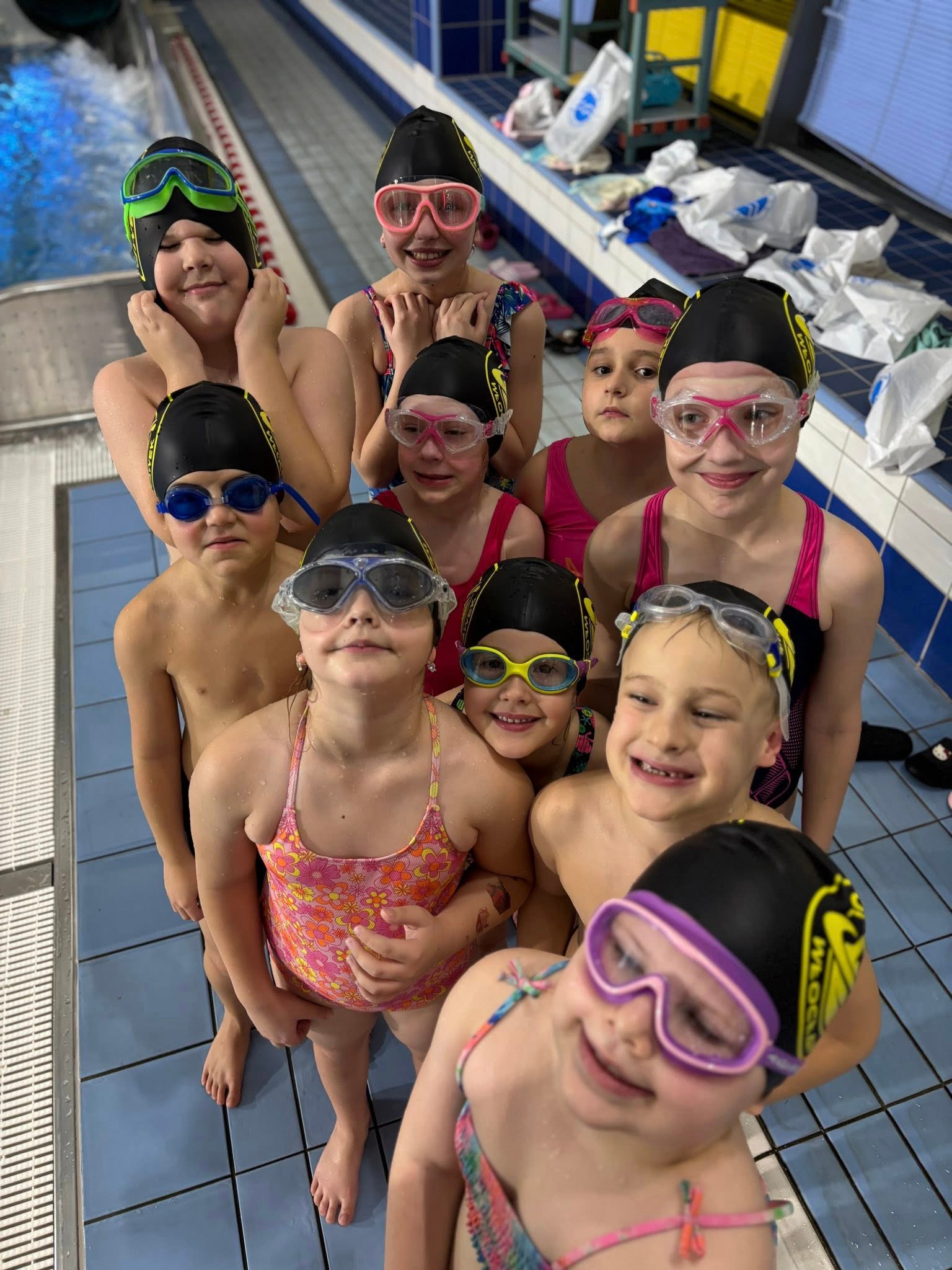 Grupa uśmiechniętych dzieci w czepkach i okularach pływackich, stojących na kafelkach przy basenie, gotowych do zajęć. Widok z góry, jasne oświetlenie.