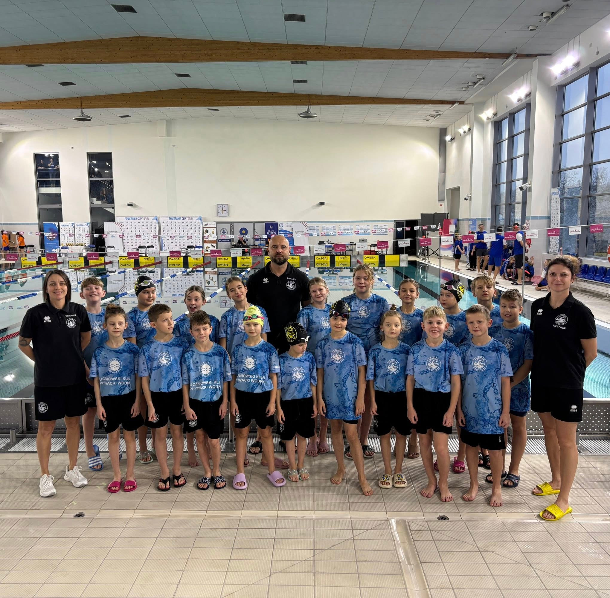 Grupa dzieci w strojach pływackich z trenerami na tle basenu. Uczniowski Klub Pływacki Wodnik Wodny. Zawody pływackie Pomerania Cup Toruń.