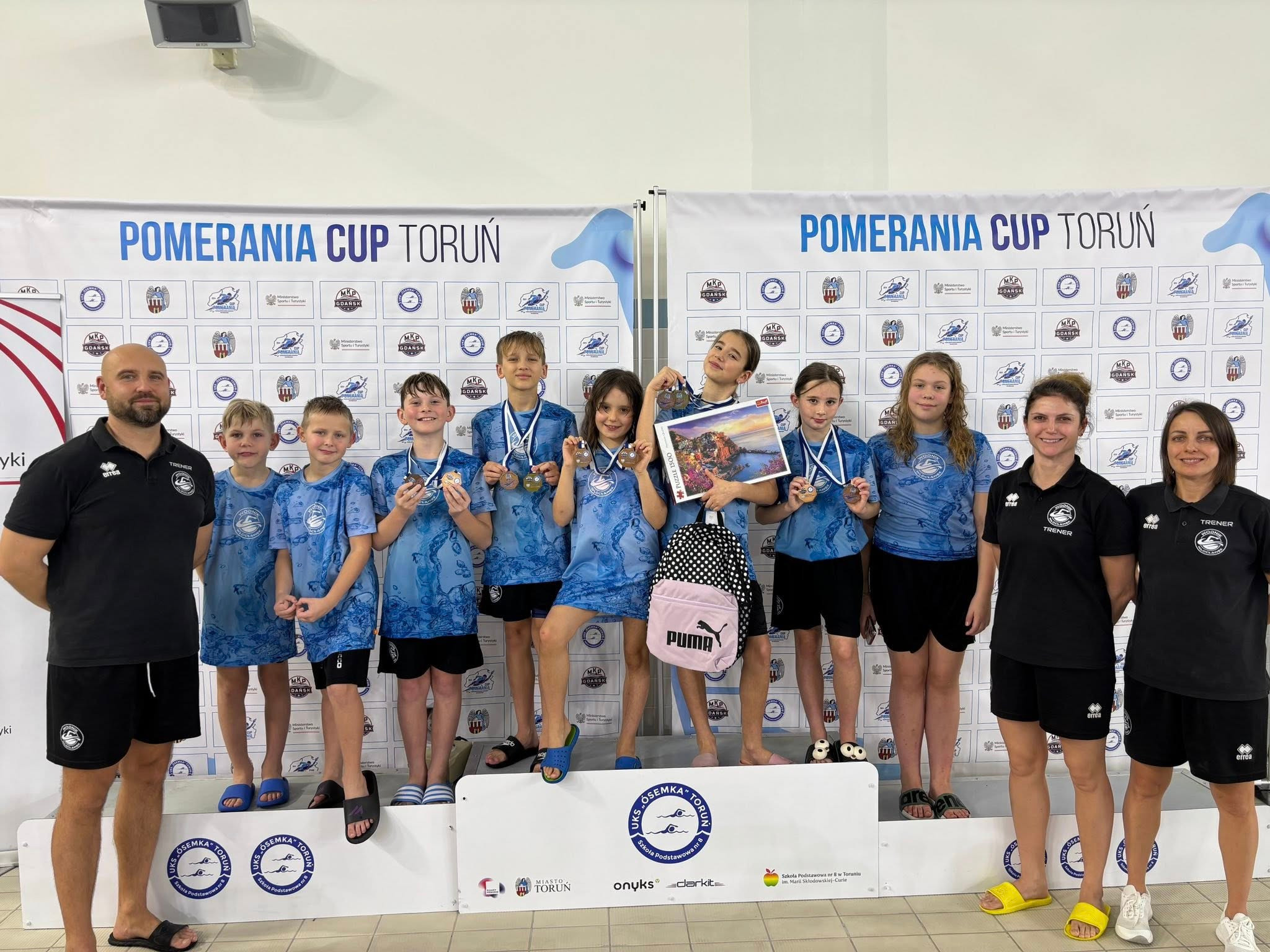 Grupa dzieci z medalami na podium po zawodach pływackich Pomerania Cup Toruń, z trenerami po bokach. W tle baner z logo zawodów i klubu.