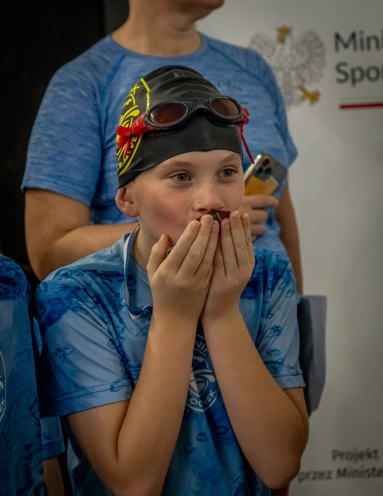 Chłopiec w czepku i okularkach pływackich je czekoladę po zawodach, trzymając ręce przy ustach. W tle osoby i logo Ministerstwa Sportu.