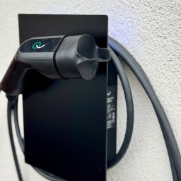 MMS System Sp. z o. o. - Czarny uchwyt z kablem i wtyczką do ładowania pojazdów elektrycznych, zamontowany na teksturowanej, jasnej ścianie. Widoczne logo na wtyczce.