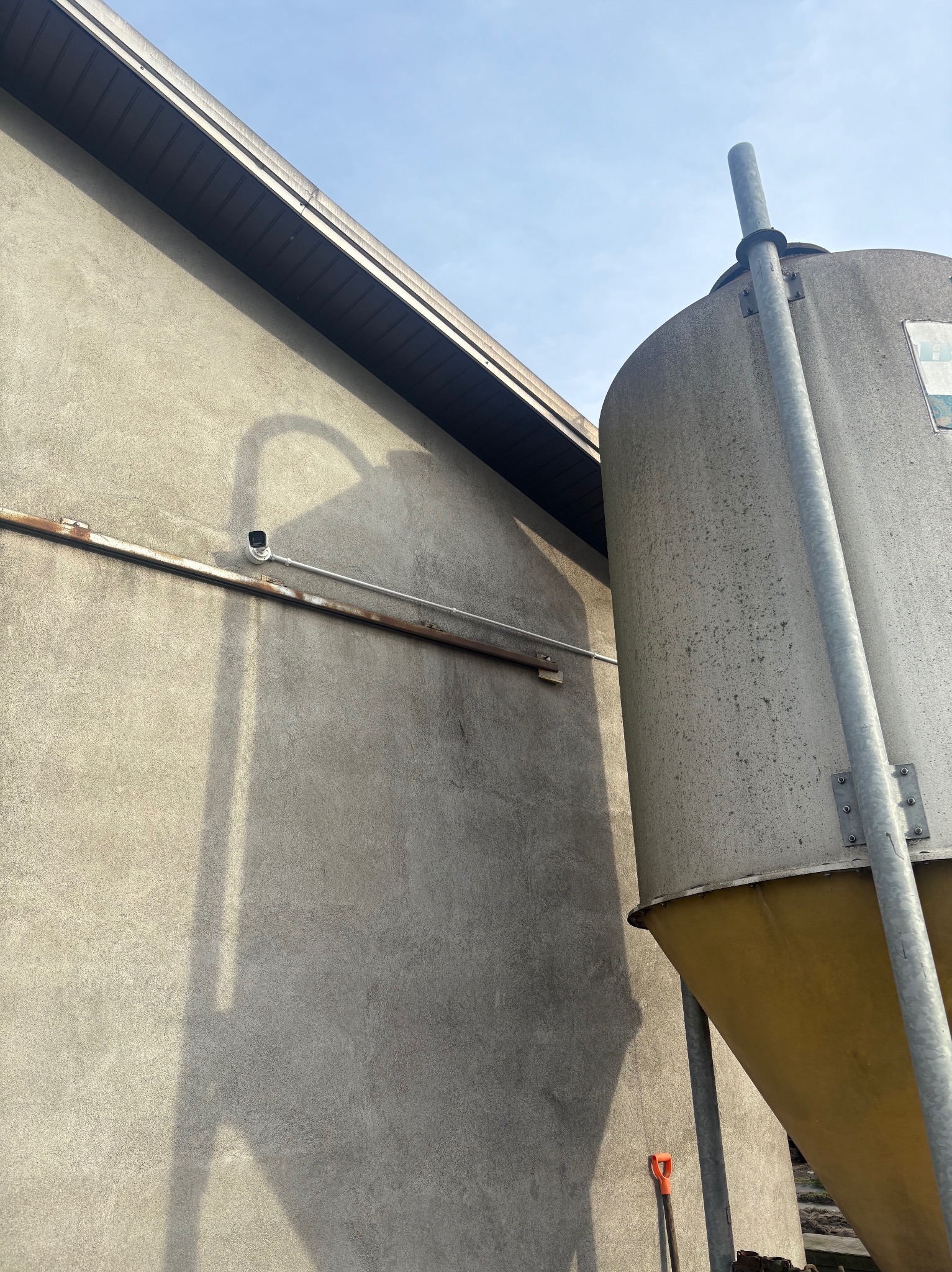 Kamera monitoringu na elewacji budynku, obok silos. Widoczne rury instalacyjne i cień. Ujęcie z dołu, w tle błękitne niebo.