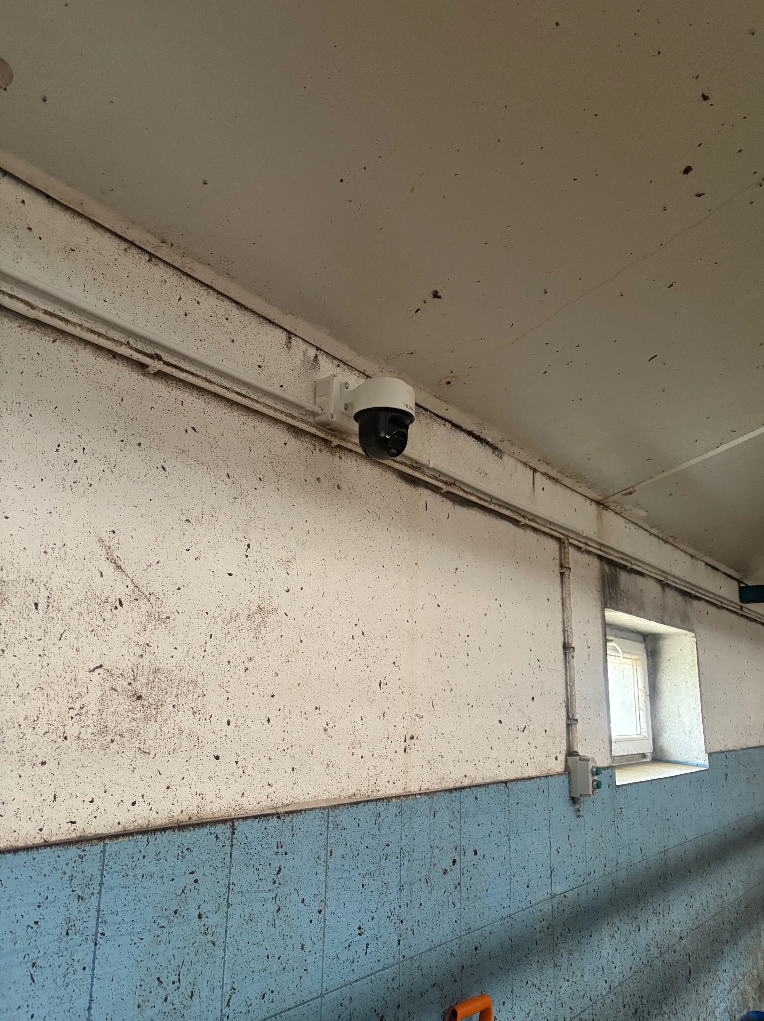 Kamera monitoringu PTZ zamontowana na suficie w pomieszczeniu z oknem i instalacją elektryczną. Widoczne ślady użytkowania na ścianach i suficie.