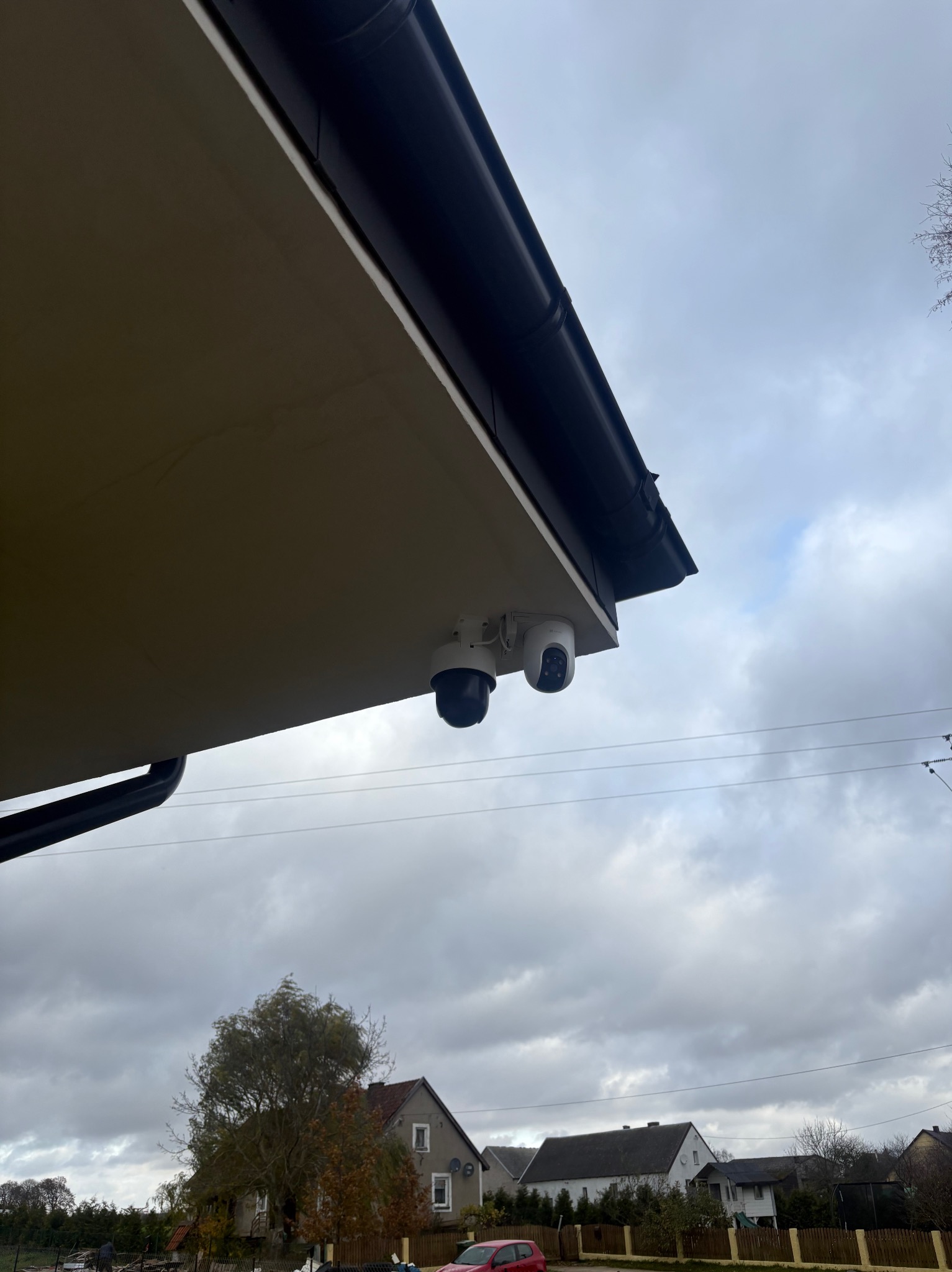 Dwie kamery monitoringu, jedna kopułkowa, druga obrotowa, zamontowane pod okapem budynku. Widok na pochmurne niebo i zabudowę jednorodzinną.