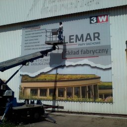 Engage Media - Montaż wielkoformatowej reklamy na elewacji w Poznaniu. Pracownik na podnośniku instaluje baner firmy Lemar, dystrybutora materiałów budowlanych, z widocznym logo 3W.