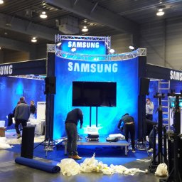 Engage Media - Montaż stoiska targowego Samsung z podświetlanym tłem i ekranami. Widoczni pracownicy podczas instalacji, elementy konstrukcyjne i sprzęt. Hala targowa w tle.