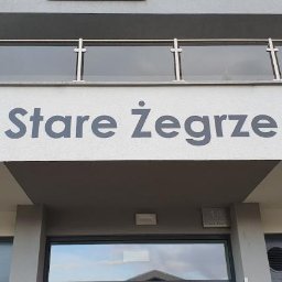 Engage Media - Szary napis 'os. Stare Żegrze 14' na elewacji budynku, nad wejściem. Widoczny balkon z szklaną balustradą i fragmenty okien. Ujęcie z dołu.