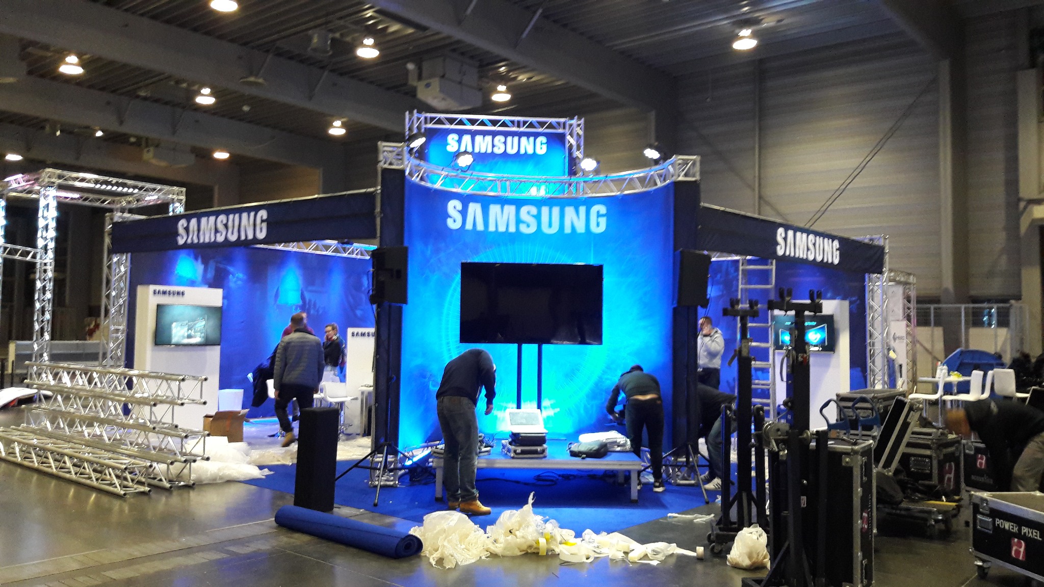 Montaż stoiska targowego Samsung z podświetlanym tłem i ekranami. Widoczni pracownicy podczas instalacji, elementy konstrukcyjne i sprzęt. Hala targowa w tle.