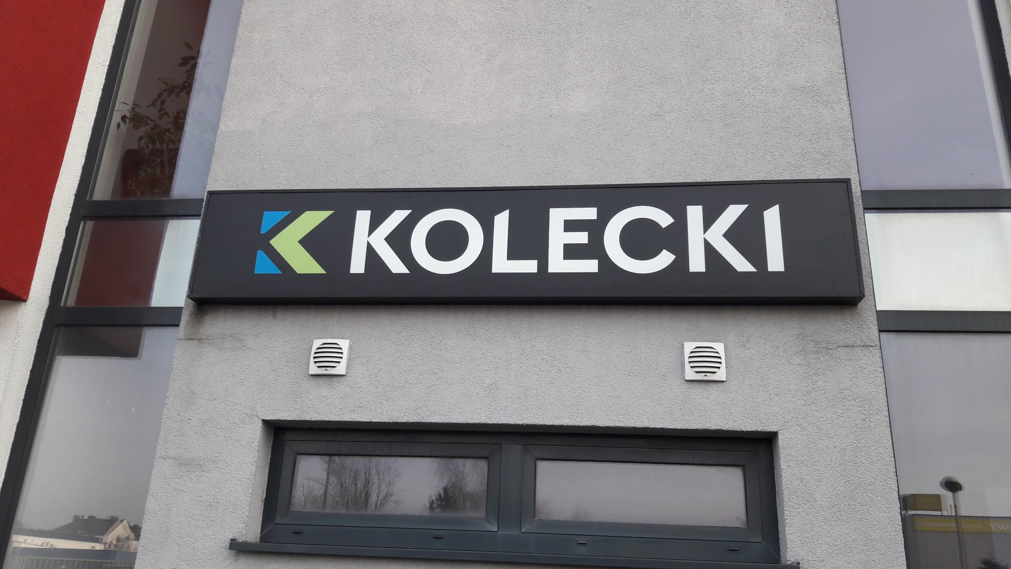 Szyld firmy 'KOLECKI' z logo w kolorach niebieskim i zielonym, umieszczony na szarej fasadzie budynku z oknami. Widoczne detale architektoniczne i faktura ściany.