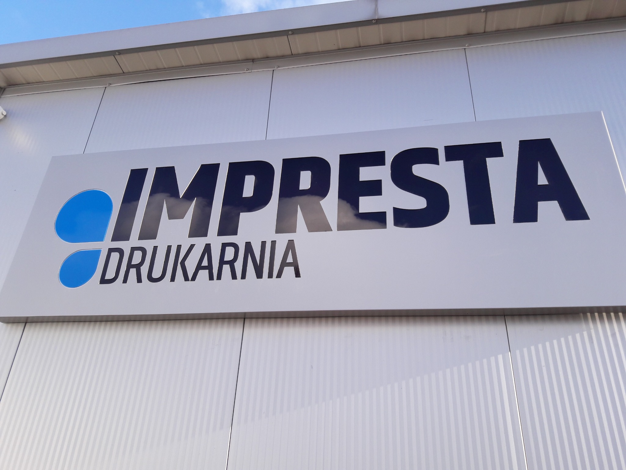 Szyld drukarni 'IMPRESTA' z niebieskim logo i czarnym napisem na białym tle, zamontowany na elewacji budynku z blachy trapezowej.