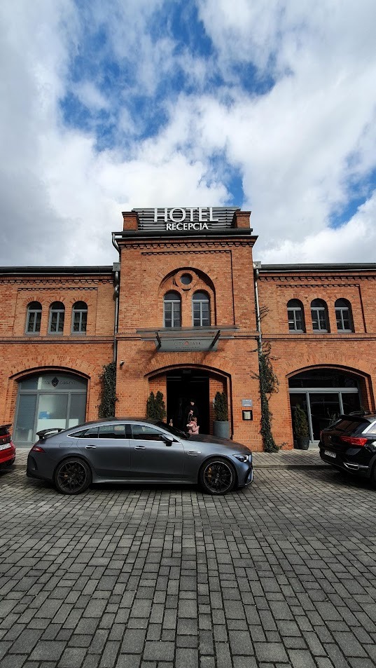 Szary Mercedes-AMG GT 63 S E Performance zaparkowany przed ceglanym budynkiem hotelu z napisem 'Hotel Recepcja' w słoneczny dzień.