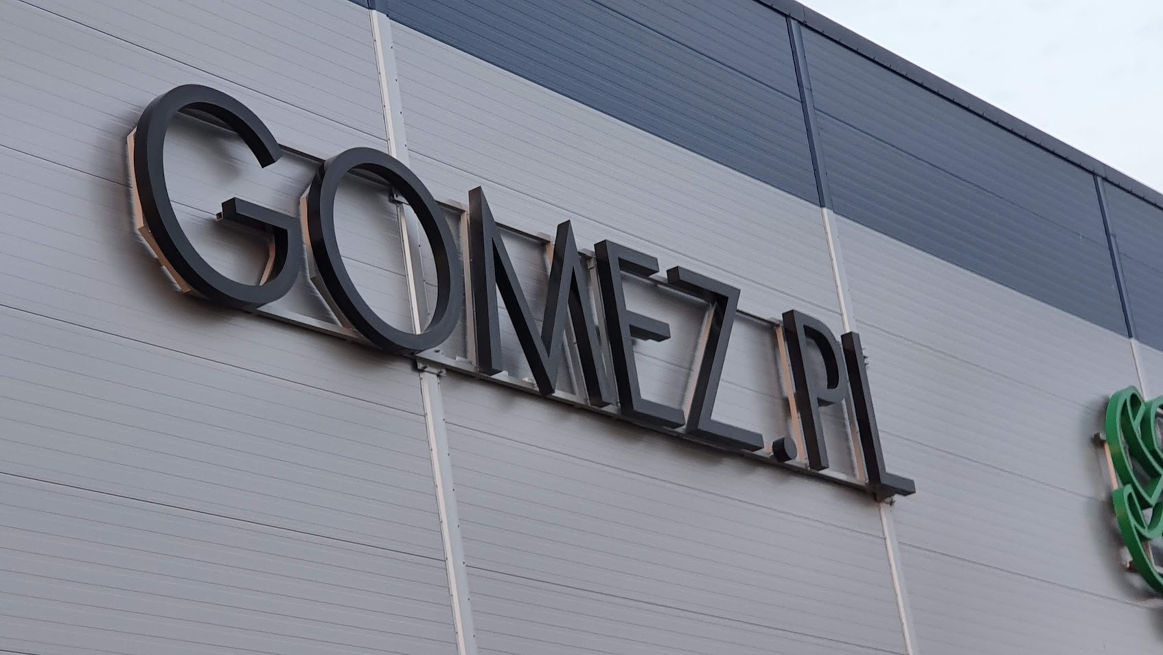 Czarny napis 'GOMEZ.PL' na szarej elewacji budynku, widoczny z dołu pod kątem. Litery przestrzenne, zamontowane na metalowej konstrukcji. Obok widoczne logo.