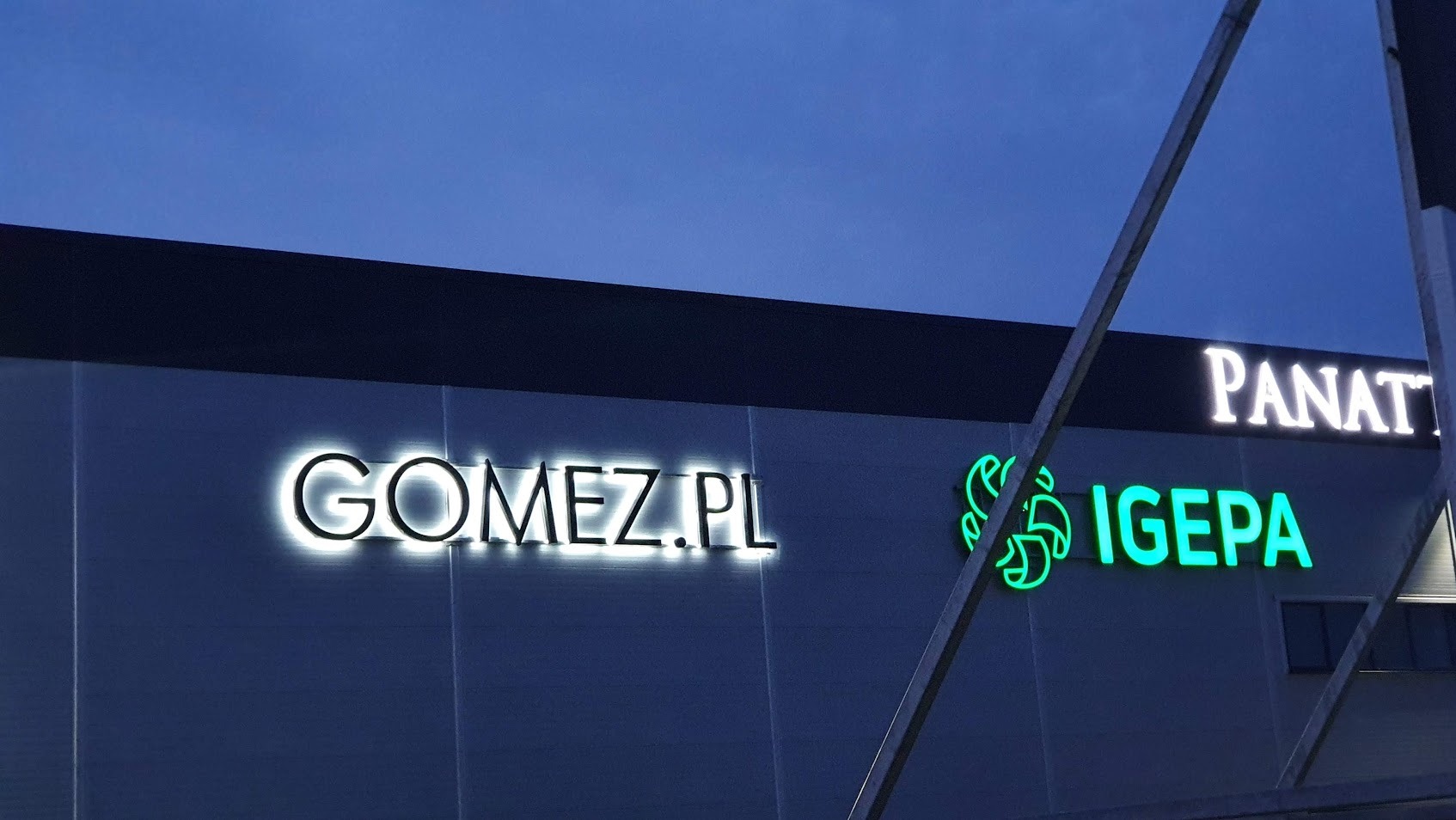 Podświetlane szyldy firmowe Gomez.pl i Igepa na elewacji budynku, widoczne wieczorem. Szyldy wykonane w różnych stylach, Gomez.pl z białą obwódką, Igepa zielony.