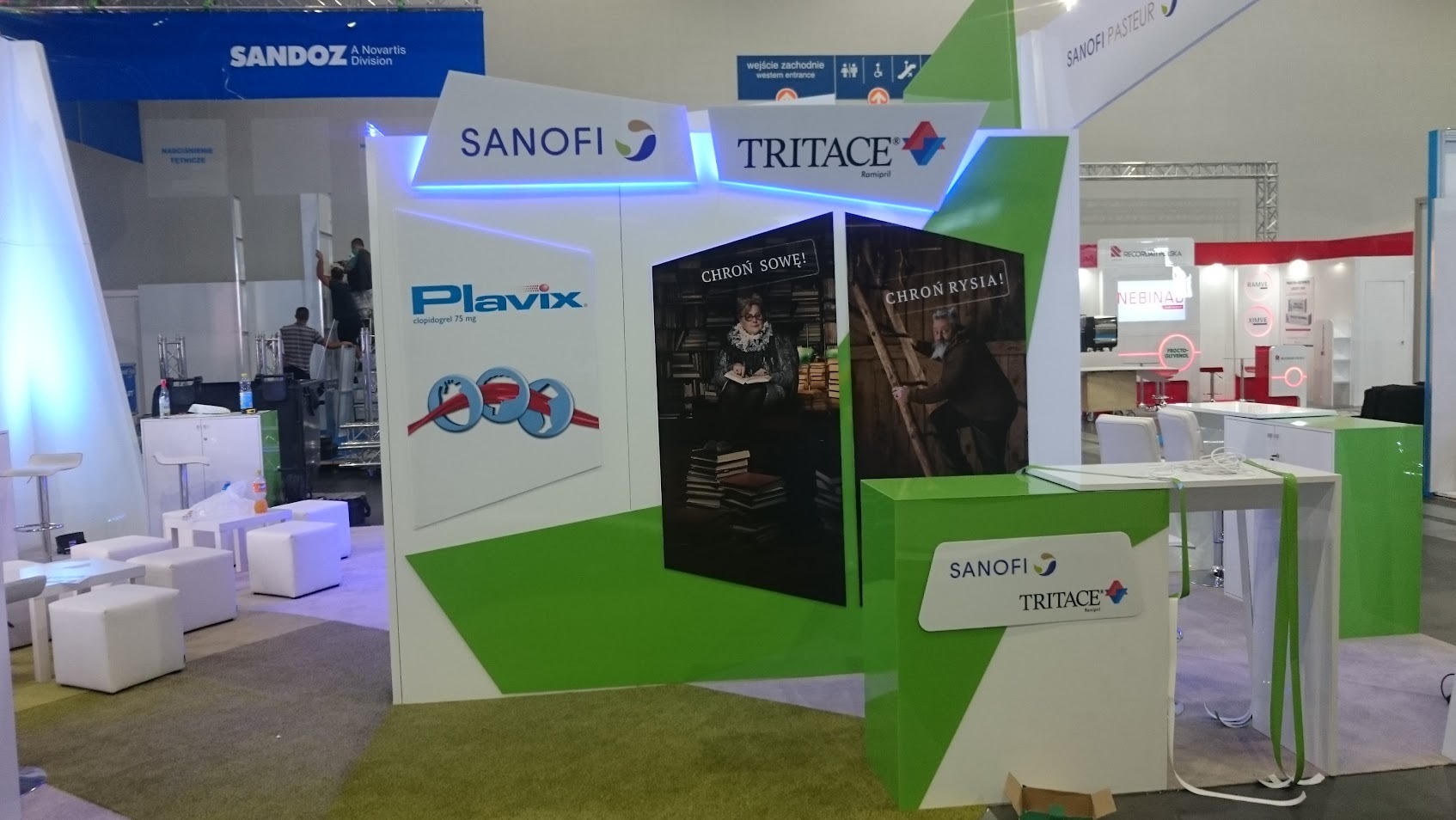Stoisko targowe Sanofi z logo Tritace i Plavix, z grafikami 'Chroń Sowę!' i 'Chroń Rysia!', oświetleniem LED i elementami w kolorze białym i zielonym.