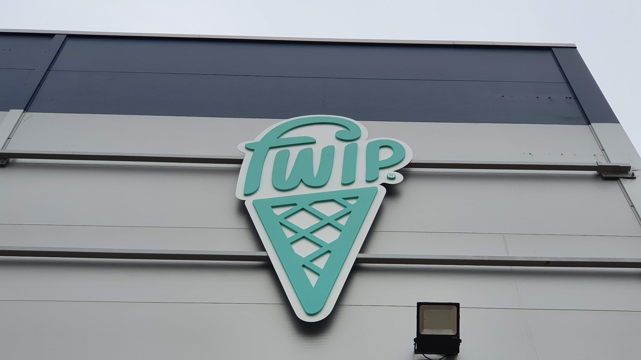Turkusowe logo 'fwip' w kształcie lodowego rożka na szarej fasadzie budynku z widocznym oświetleniem zewnętrznym.